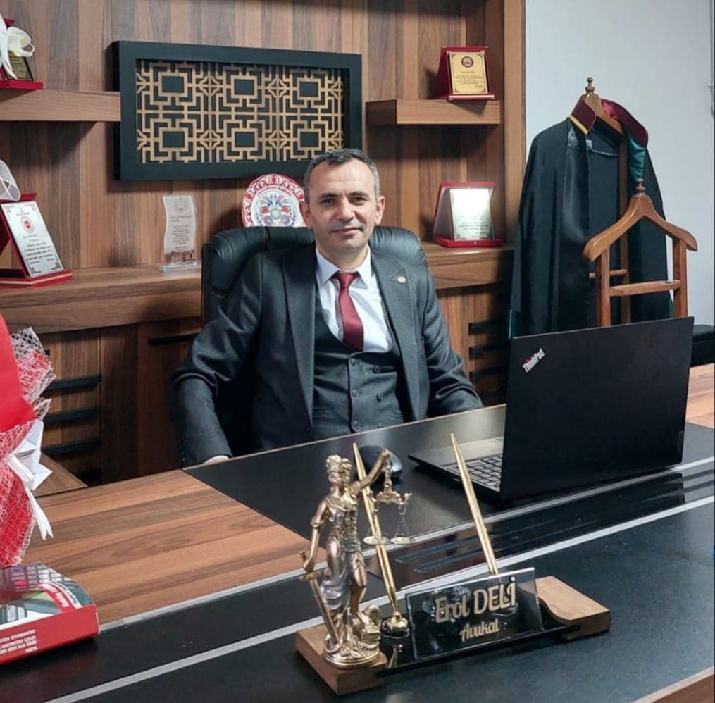 Attorney Erol Deli at his desk in the Bahçelievler office, with a lawyer’s robe and plaques behind him and a statue of Justice and a nameplate reading “Erol Deli, Attorney” on the desk.